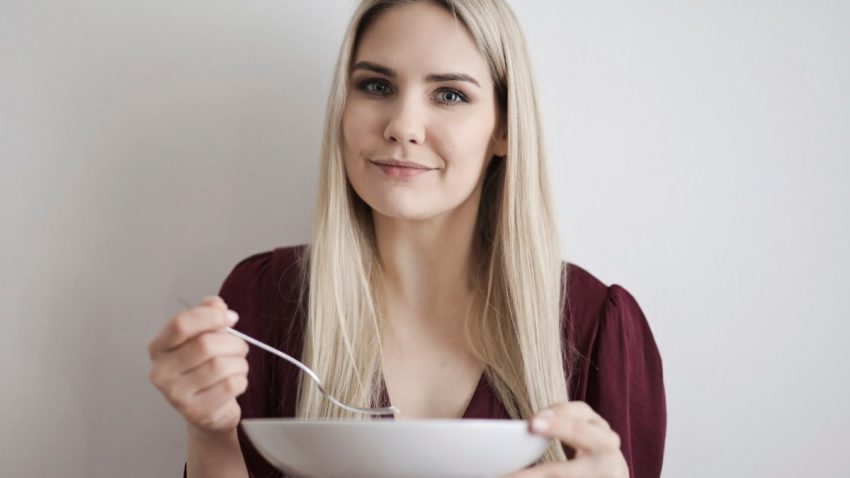 Smiling woman with blonde hair in a maroon top holding a bowl against a white background.